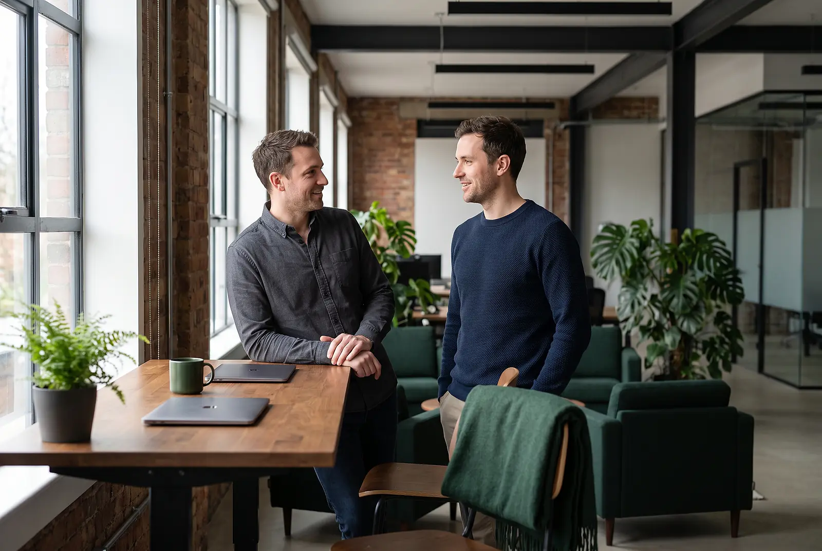 Zwei IT-Experten unterhalten sich an einem Stehtisch im modernen Loft-Büro über Mitarbeiter-Benefits